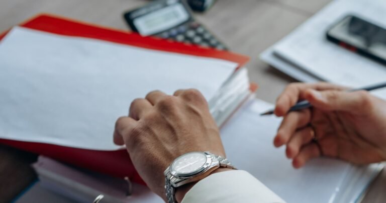 Bàn tay đeo đồng hồ đặt trên tập hồ sơ giấy tờ cùng máy tính và điện thoại trên bàn làm việc