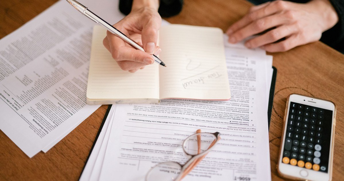 Bàn tay đang ghi chép vào sổ tay trên bàn gỗ đầy giấy tờ thuế, kính mắt và điện thoại