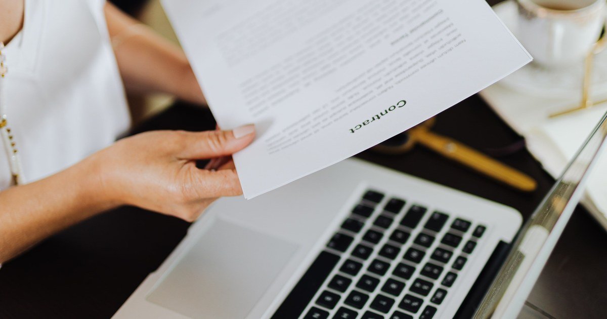 Bàn tay cầm tờ giấy ghi chữ Contract phía trên laptop