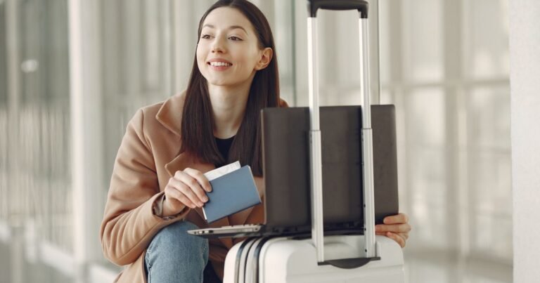 Người phụ nữ ngồi tại sân bay, tay cầm hộ chiếu, bên cạnh vali kéo và laptop