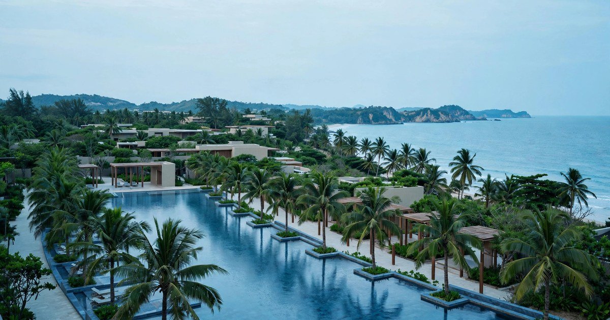 Khu resort ven biển với hồ bơi dài, hàng dừa dọc lối đi, vịnh biển và núi đá ở phía xa.