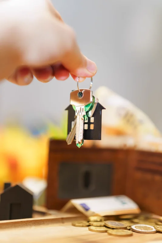 Hand holding house keys with house keychain and coins; no people visible