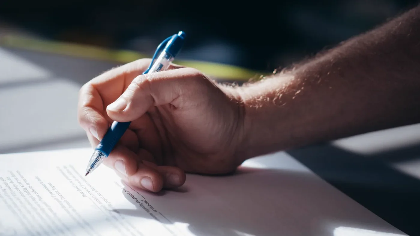 Hand signing a legal document with a blue pen; no face visible