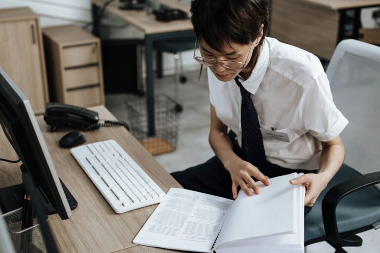 Professional in office attire reviewing documents at a desk with computer and book
