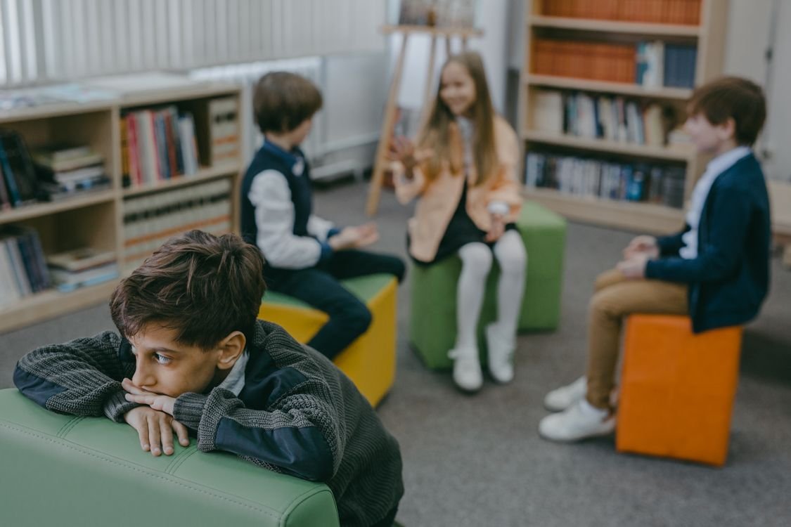 A sad child isolated in a school library while peers chat in the background