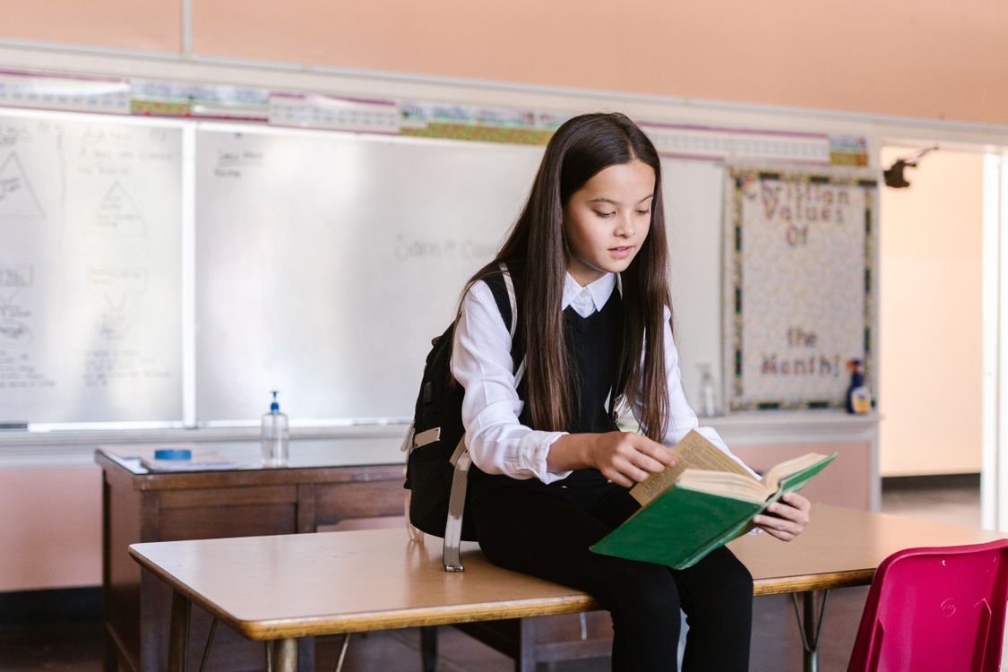 孩子在越南上学必须会越南语吗? 1 Young girl sitting on a desk in a classroom reading a book wearing a backpack