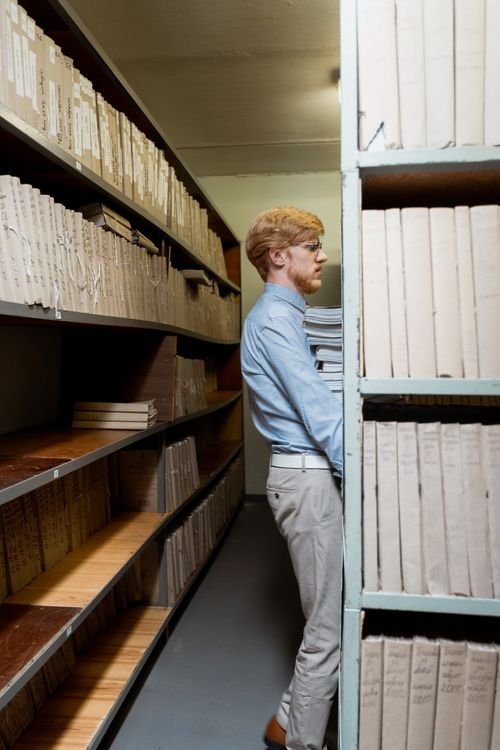 Side view of a man organizing documents in a dimly lit archive room with shelves of files