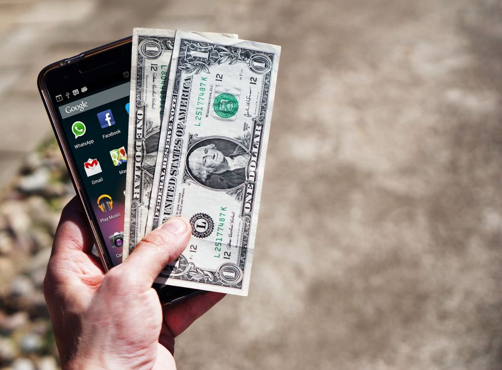 Close-up of a hand holding US dollar bills and a smartphone outdoors