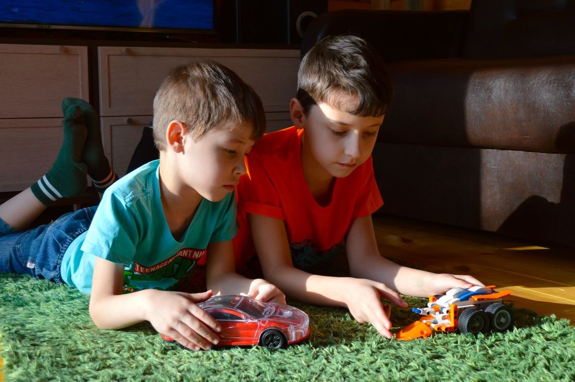 Two young boys lying on a green carpet playing with toy cars indoors