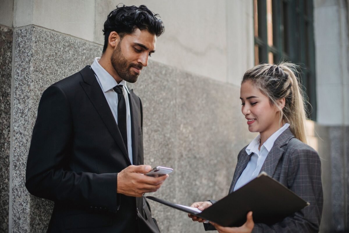 Two business professionals engaged in conversation outside a building, holding a smartphone and documents.